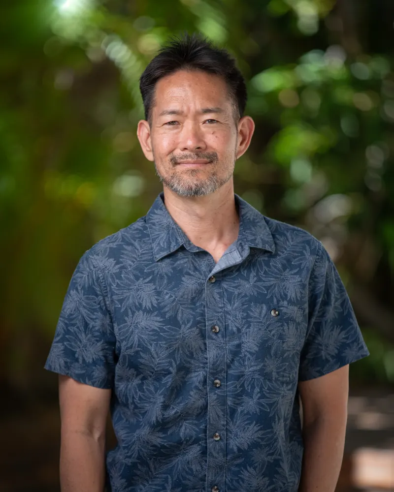 Dave Kusumoto in a navy aloha shirt, in front of tropical foliage