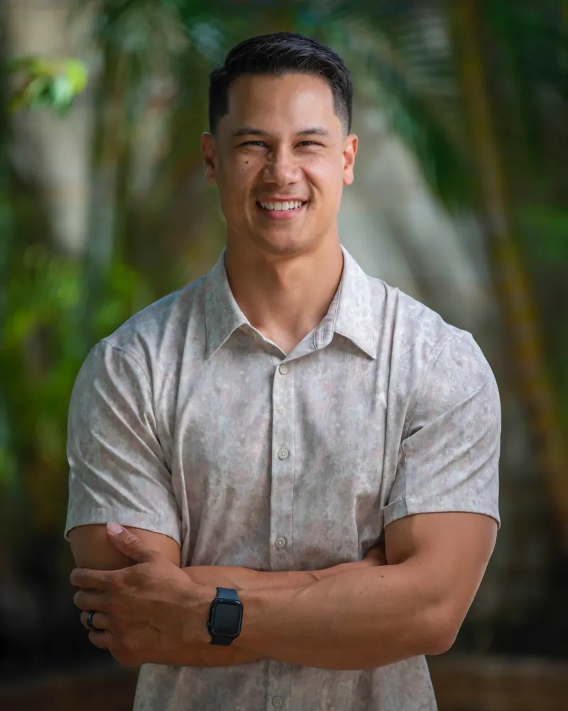 Kawika Lopez, smiling, arms crossed, in front of tropical foliage